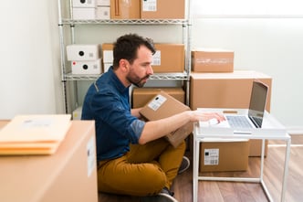 hispanic-young-man-sitting-his-office-floor-while-holding-package-typing-laptop-check-customer-order-from-online-shop
